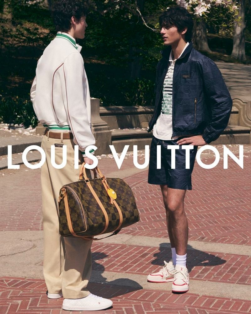Mathieu Simoneau and Akbar Shamji stand on a brick park path in Central Park, with Simoneau in a cream and green track jacket and tan chinos holding a Damier Keepall, and Shamji in a navy quilted bomber over a striped tee and matching shorts