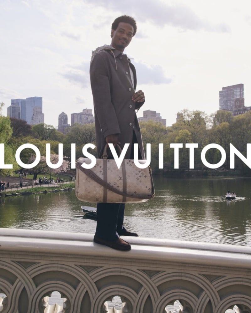 Tyshawn Jones stands on the stone railing of Bow Bridge in Central Park wearing a long taupe car coat over black trousers and brown loafers, holding a Monogram Keepall against the Manhattan skyline