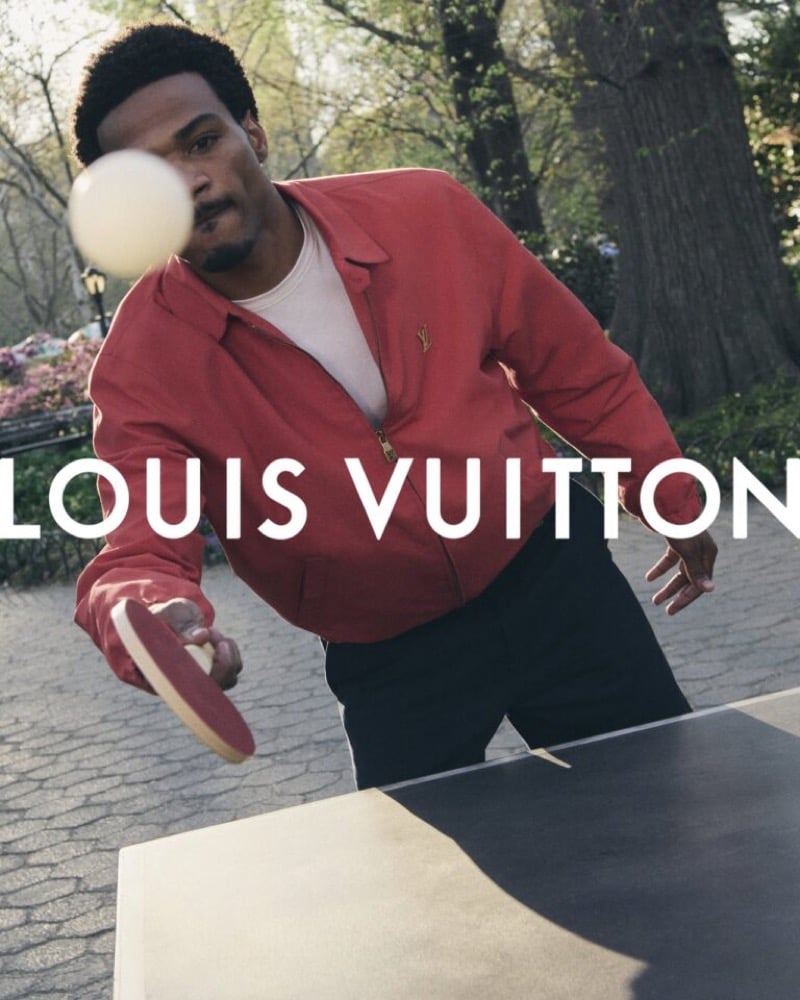 Tyshawn Jones reaches for a ping-pong ball mid-volley on a Central Park table tennis court, wearing the red LV-monogrammed harrington jacket over a white tee with black trousers