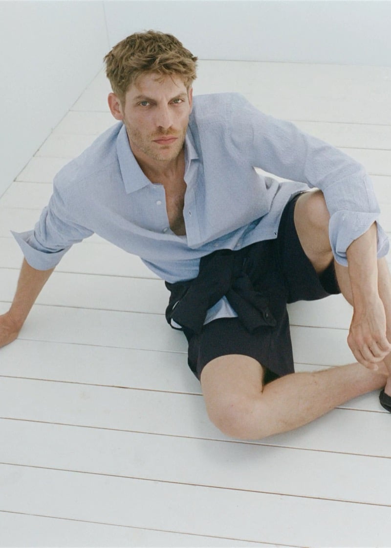 Baptiste Radufe lounges on a white plank floor in a light blue linen shirt and dark shorts for Mango Man summer 2026