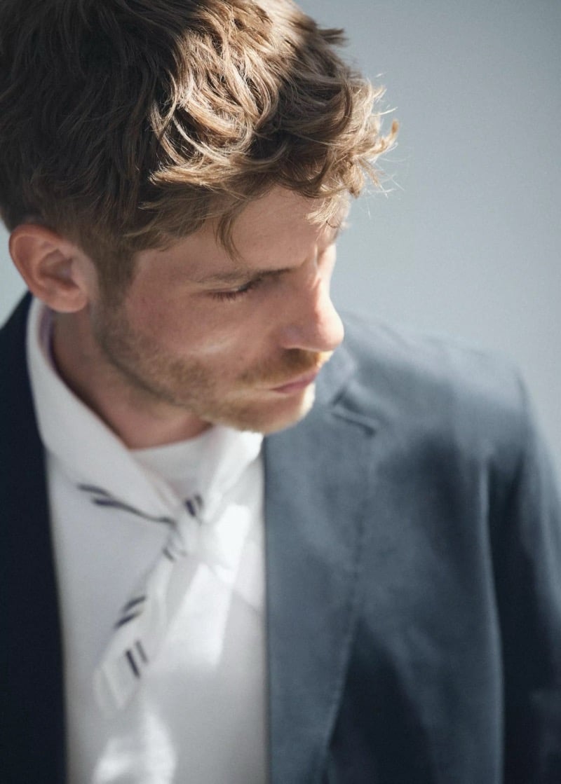Close-up of Baptiste Radufe in a slate blue cotton blazer over a white shirt with a striped silk neckerchief knotted at the collar for Mango Man summer 2026