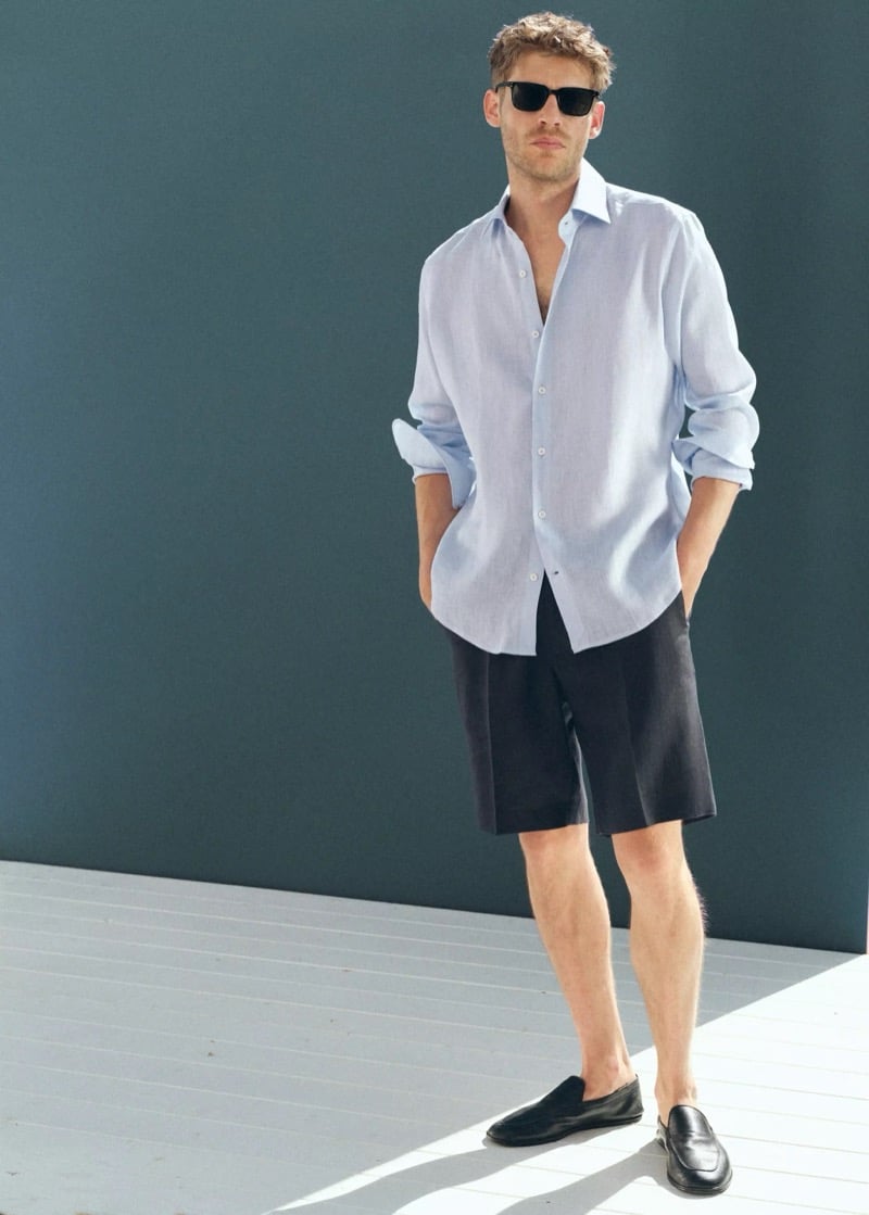 Baptiste Radufe wears a light blue linen shirt with charcoal tailored Bermuda shorts, black sunglasses, and black mules for Mango Man summer 2026