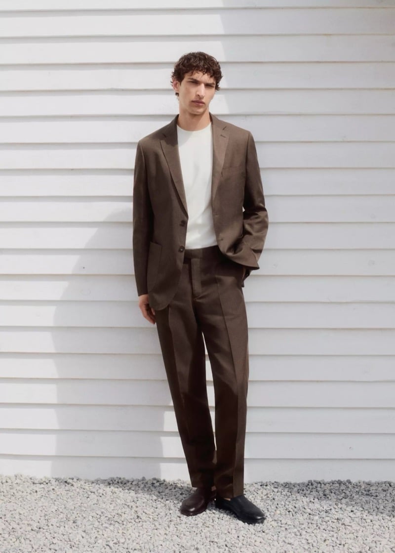 Habib Masovic in a chocolate brown lyocell-linen single-breasted suit worn over a white crewneck T-shirt and black leather loafers, standing on pale gravel against a white clapboard wall