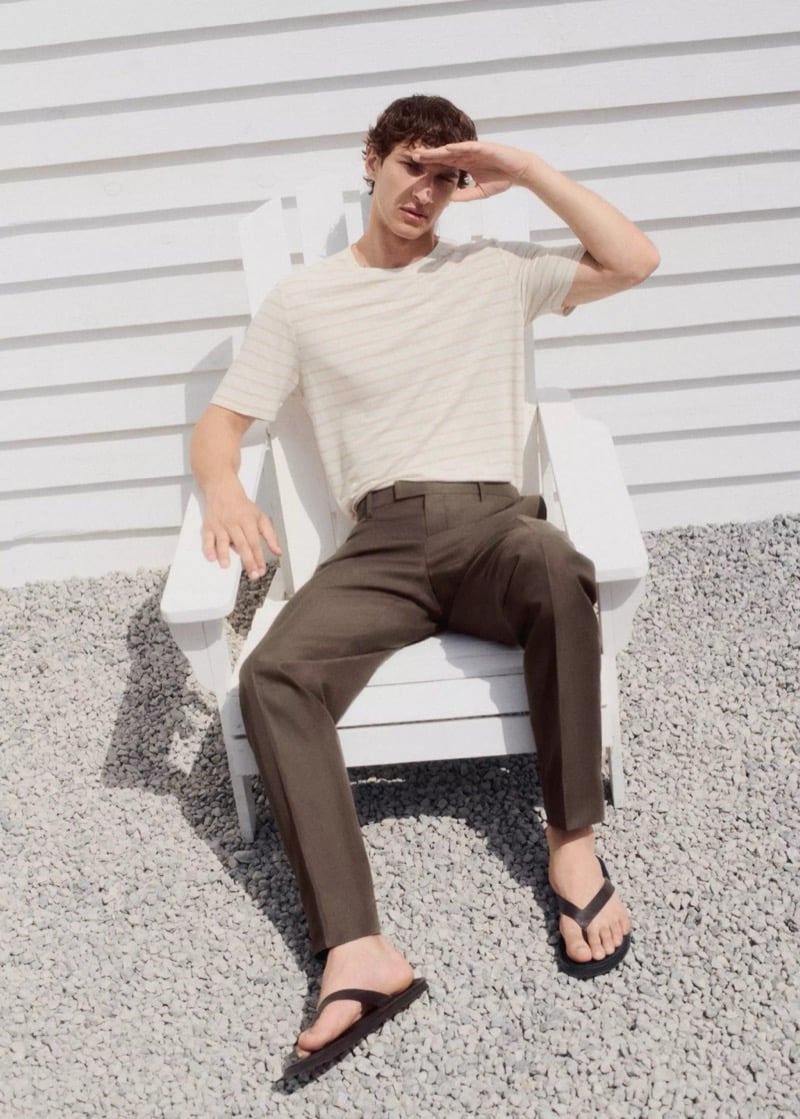 Habib Masovic reclining in a white Adirondack chair in a cream striped short-sleeve T-shirt, olive-brown pleated trousers, and black leather flip flops, shading his eyes from the sun
