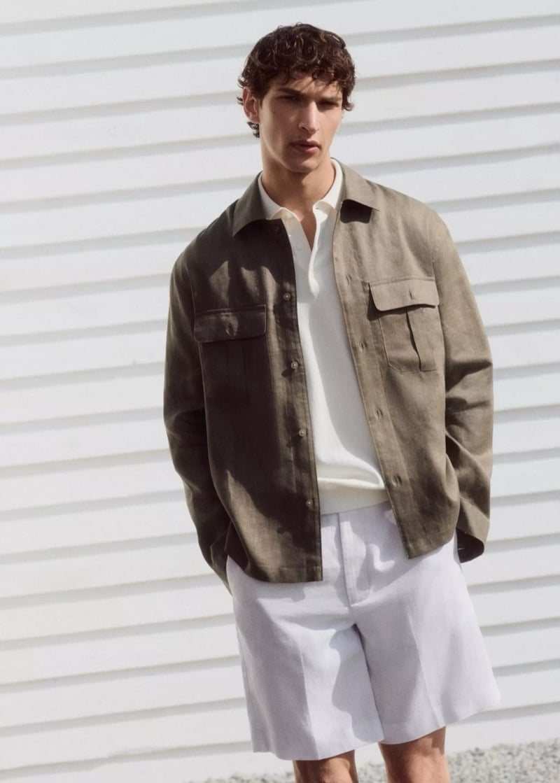 Habib Masovic in an olive linen overshirt with twin chest pockets layered over a cream knit polo and off-white linen Bermuda shorts, photographed against a white clapboard wall