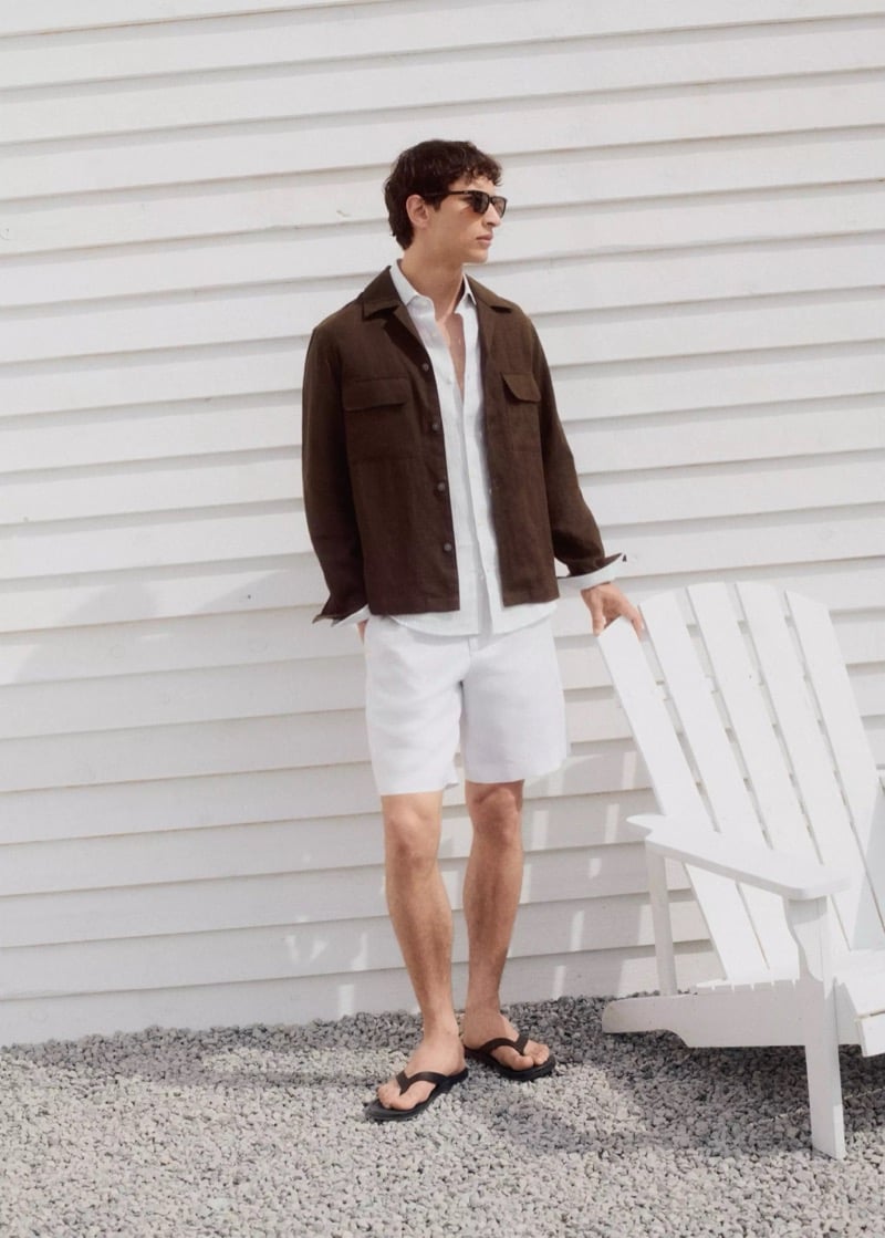 Habib Masovic in dark sunglasses, a chocolate brown linen overshirt unbuttoned over a pale shirt, white linen Bermuda shorts, and black leather flip flops beside a white Adirondack chair