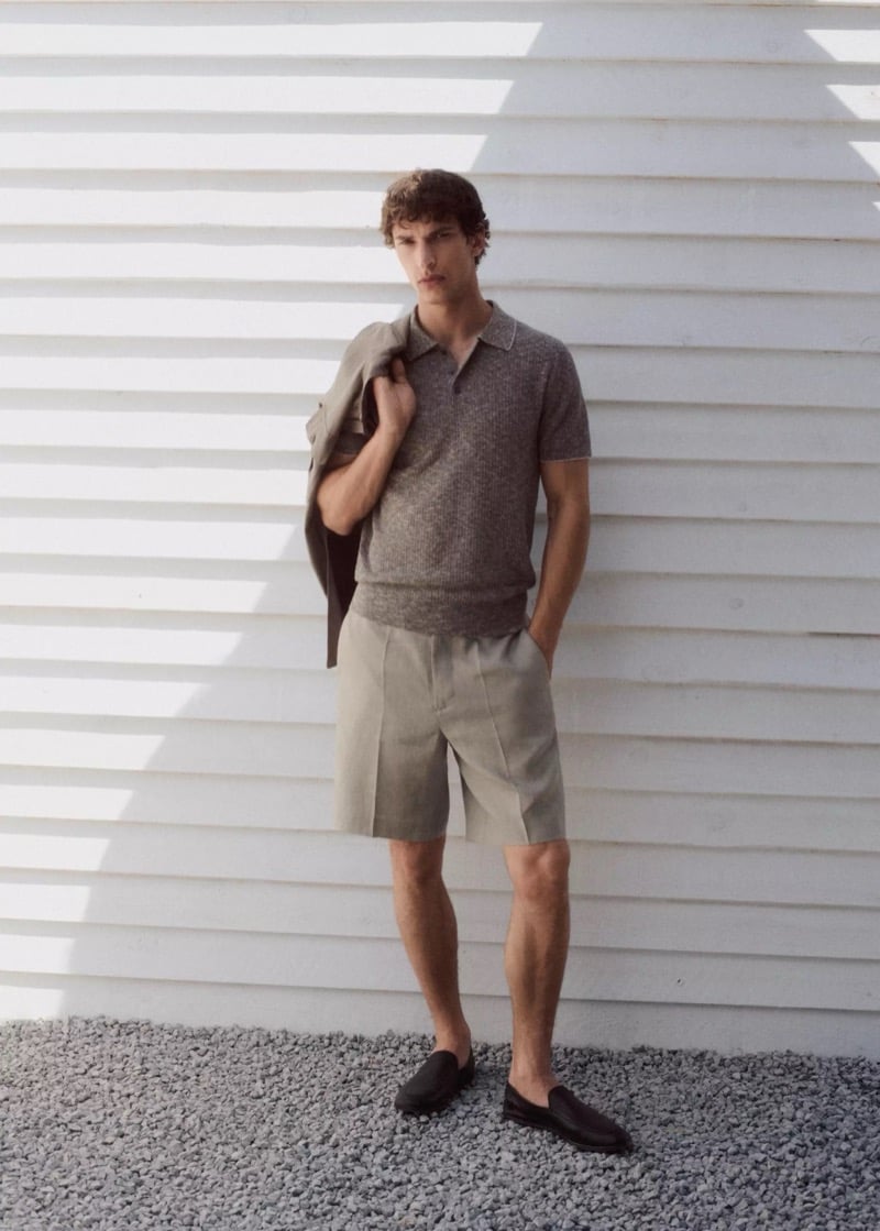 Habib Masovic with a tonal jacket draped over his shoulder, wearing a heather brown short-sleeve knit polo, sand pleated Bermuda shorts, and dark brown leather loafers on gravel
