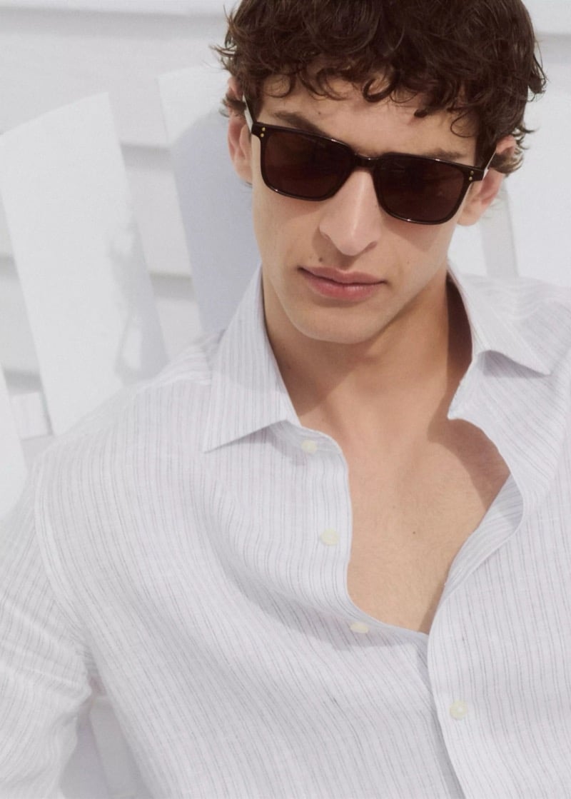 Close-up of Habib Masovic in rectangular sunglasses and a white linen shirt with fine gray pinstripes, partially unbuttoned, near a white Adirondack chair