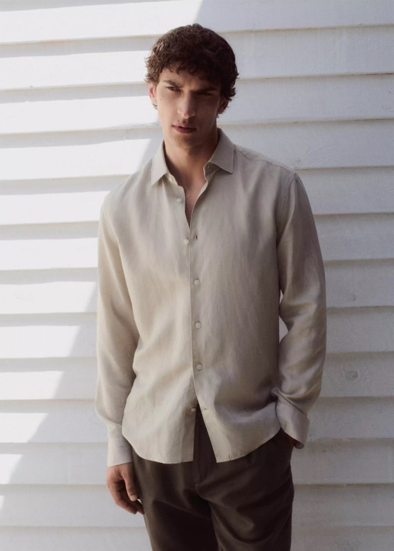 Habib Masovic in an oatmeal-toned long-sleeve linen shirt with rolled cuffs and dark chocolate trousers, standing against a sunlit white clapboard wall