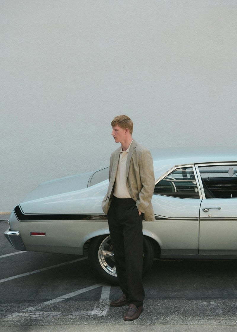 Connor Newall in profile, wearing a tan linen blazer, cream polo, black tailored trousers, and brown leather loafers