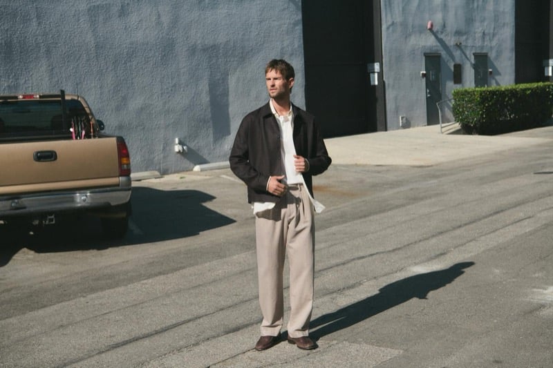 Jimmy Brighton standing in a sunlit Hollywood backlot in a dark harrington jacket over an open white shirt, beige pleated wide-leg trousers, and brown suede loafers