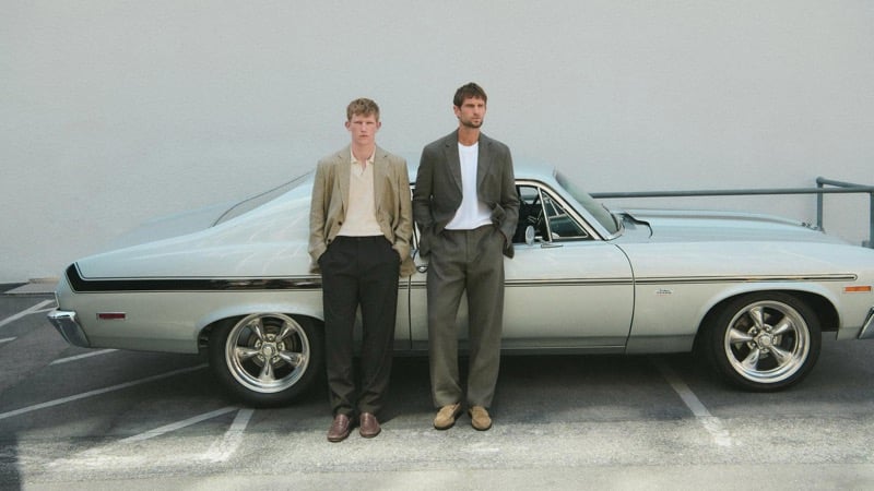 Connor Newall and Jimmy Brighton standing in front of a vintage silver muscle car in a Los Angeles parking lot, Connor in a tan linen blazer over a cream polo with black tailored trousers and brown leather loafers, Jimmy in a charcoal blazer over a white t-shirt with gray pleated trousers and tan suede loafers