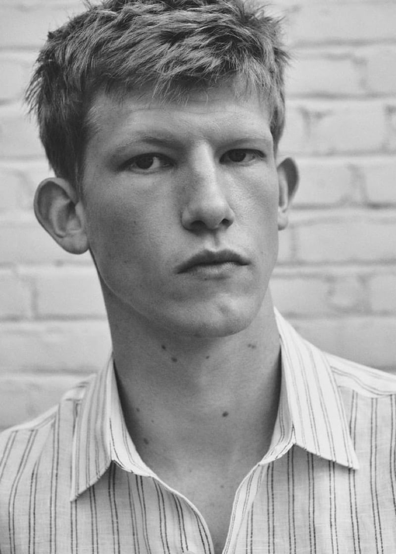 Black-and-white headshot of Connor Newall against a white brick wall in a white linen shirt finely striped with thin navy lines, collar open