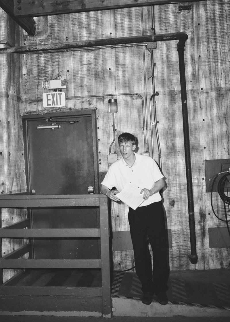 Black-and-white image of Connor Newall holding a script outside an industrial exit door in a white short-sleeve button-up and dark trousers, an EXIT sign glowing above