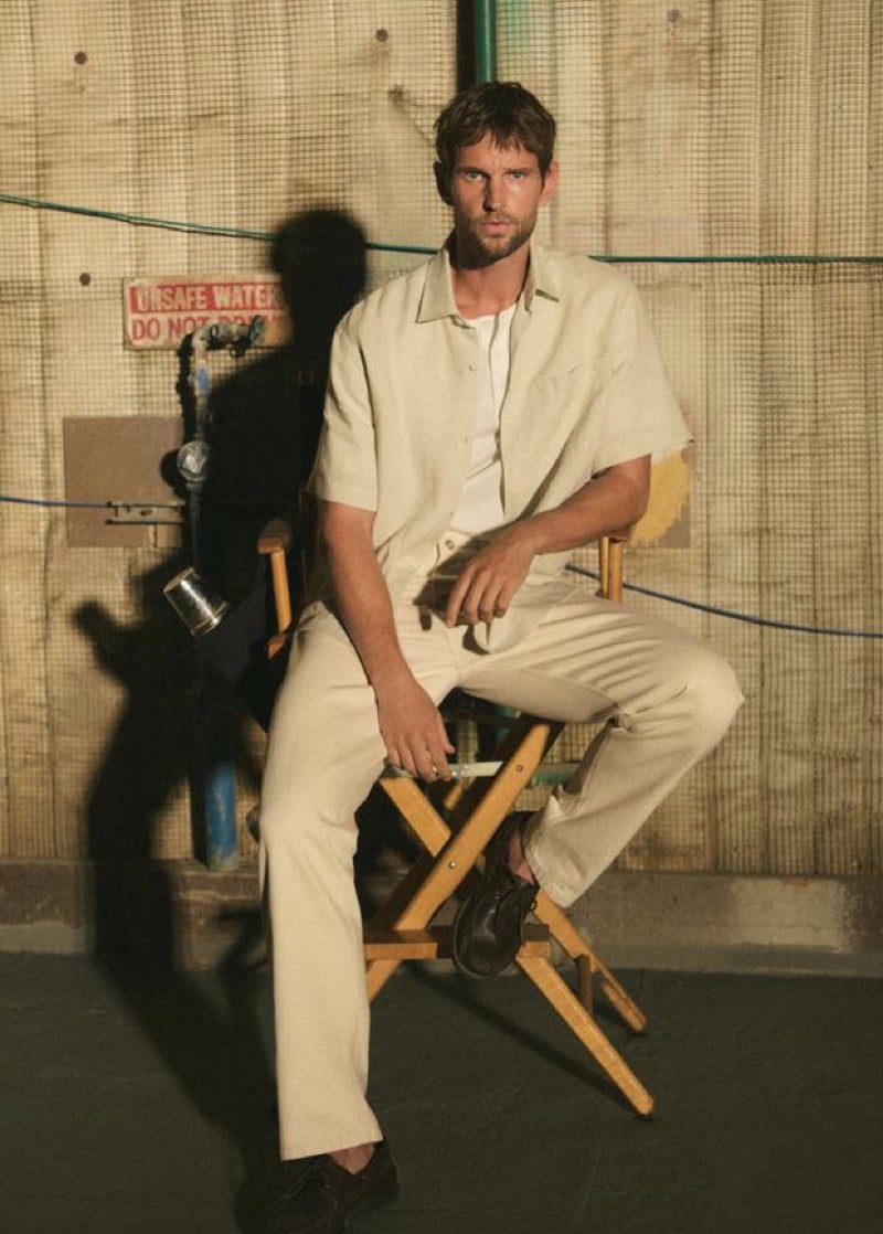 Jimmy Brighton seated on a wooden director's chair on set in a beige short-sleeve camp shirt over a white tank top, matching beige trousers, and dark brown derby shoes