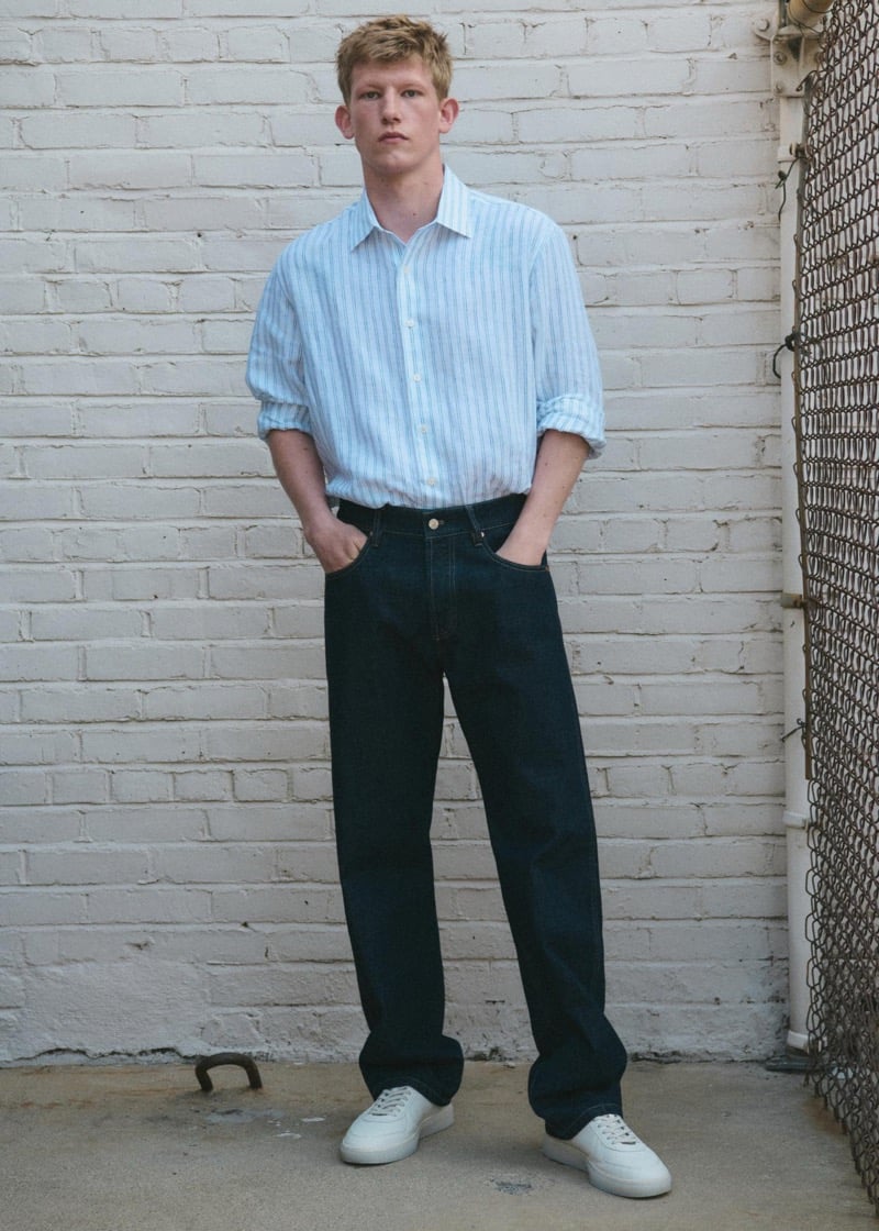 Connor Newall standing against a whitewashed brick wall in a pale blue and white striped long-sleeve linen button-up shirt, dark indigo straight-leg jeans, and white leather sneakers