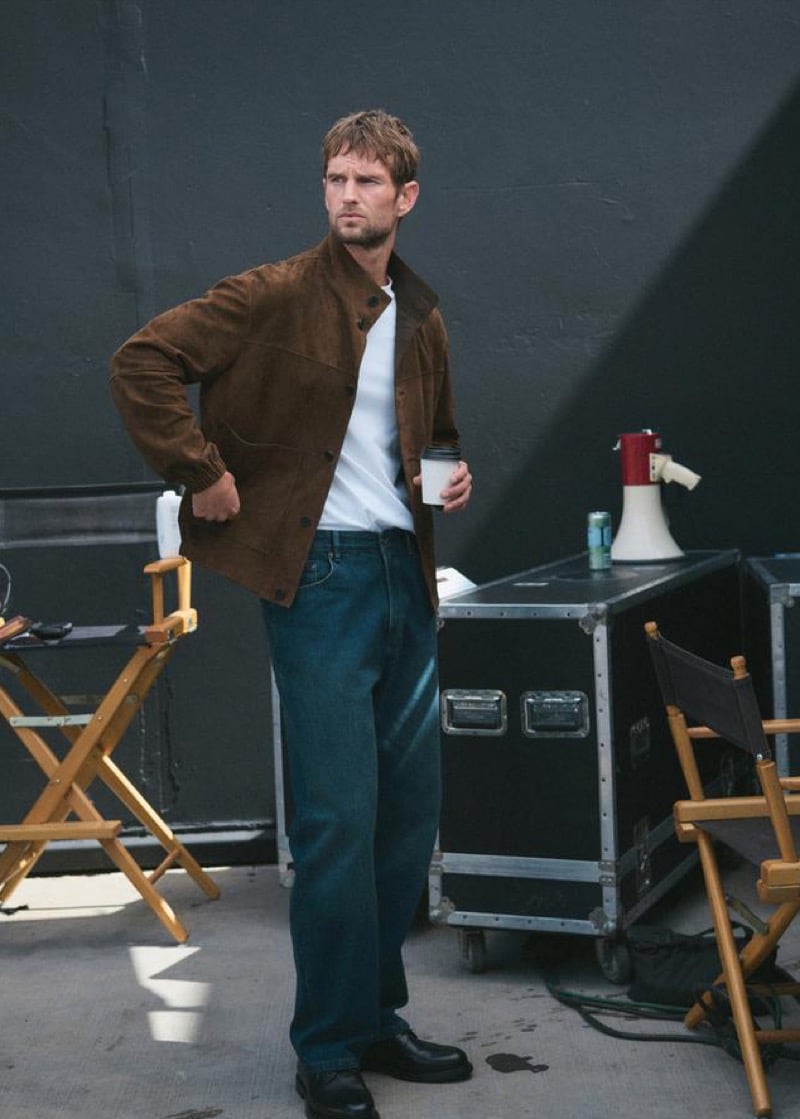 Jimmy Brighton holding a paper coffee cup backstage on a Hollywood set in a chocolate brown suede shirt jacket over a white t-shirt, dark blue straight-leg jeans, and black leather loafers, with wooden director's chairs and a red megaphone behind him