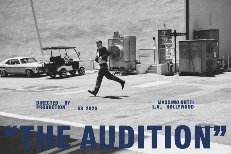 Black-and-white Massimo Dutti The Audition title card with Connor Newall mid-run across a Hollywood parking lot in a dark suit, golf carts and a vintage car parked along a studio wall