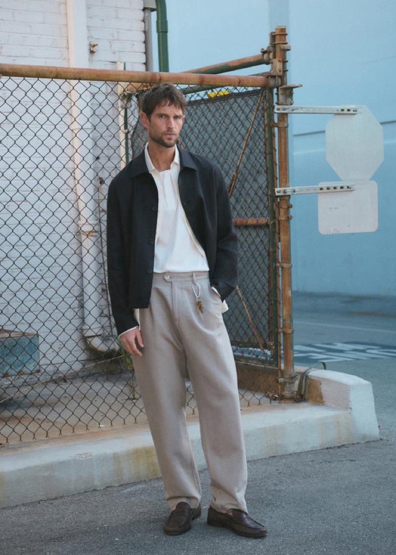 Jimmy Brighton in front of a chain link fence in a harrington jacket layered over a white shirt, beige pleated wide-leg trousers, and dark brown leather loafers
