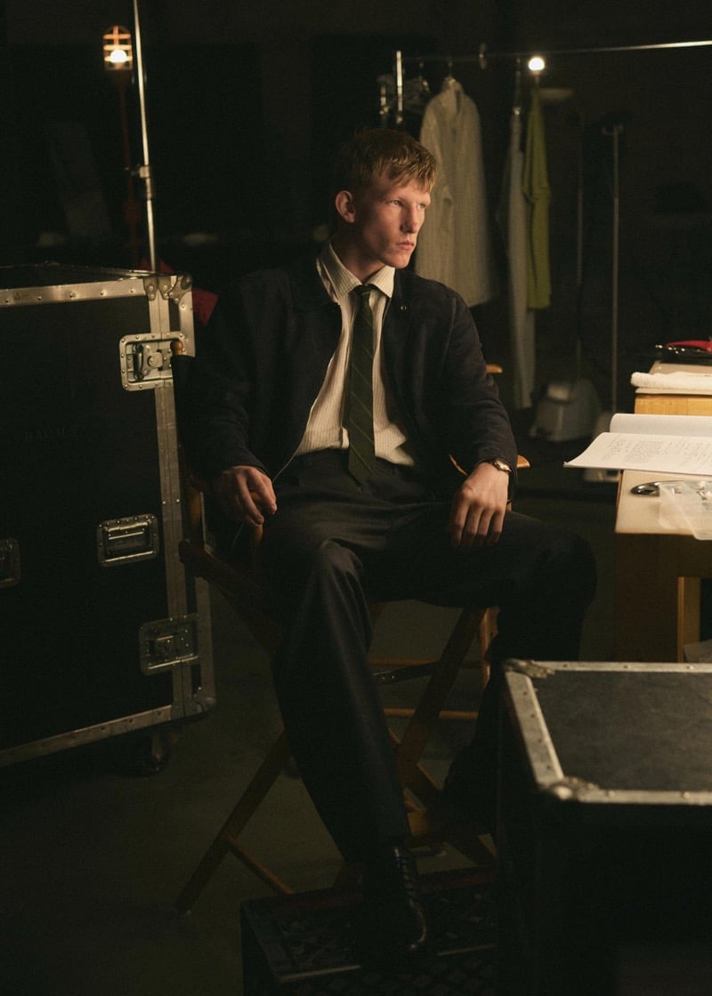 Connor Newall seated on a wooden director's chair backstage in a jacket over a white striped shirt and dark striped tie, dark trousers, and black lace-up boots, beside a wardrobe rack and equipment trunks
