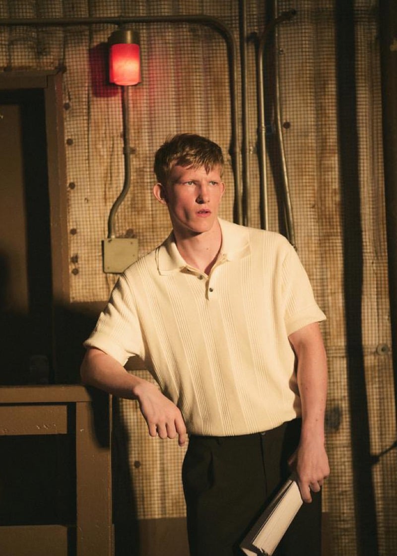 Connor Newall holding a script in a dim industrial space lit by a red wall lamp, wearing a cream ribbed knit short-sleeve polo and dark trousers