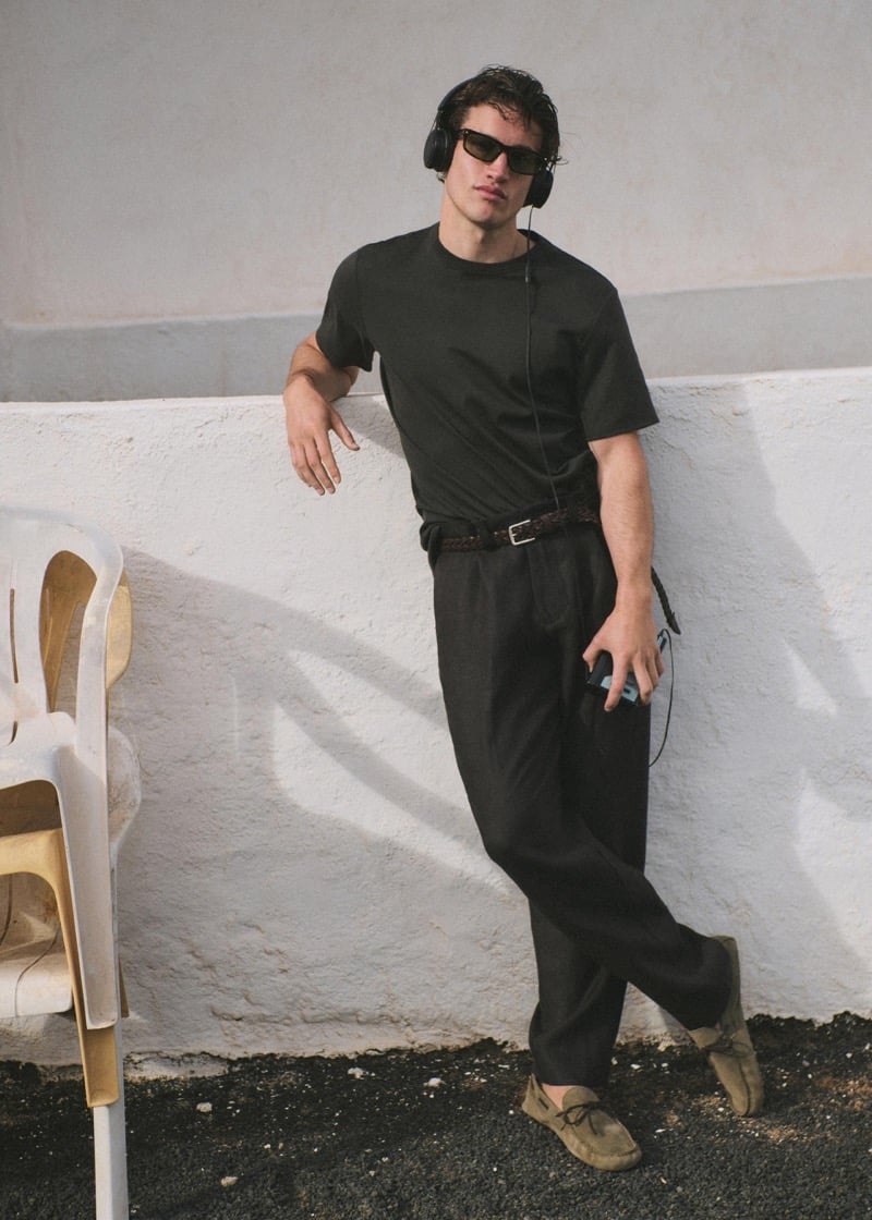 Max Knott leaning against a whitewashed wall beside a stack of plastic chairs, wearing a dark crewneck t-shirt, dark linen trousers with a braided leather belt, tan suede moccasins, over-ear headphones and tortoise sunglasses, holding a phone