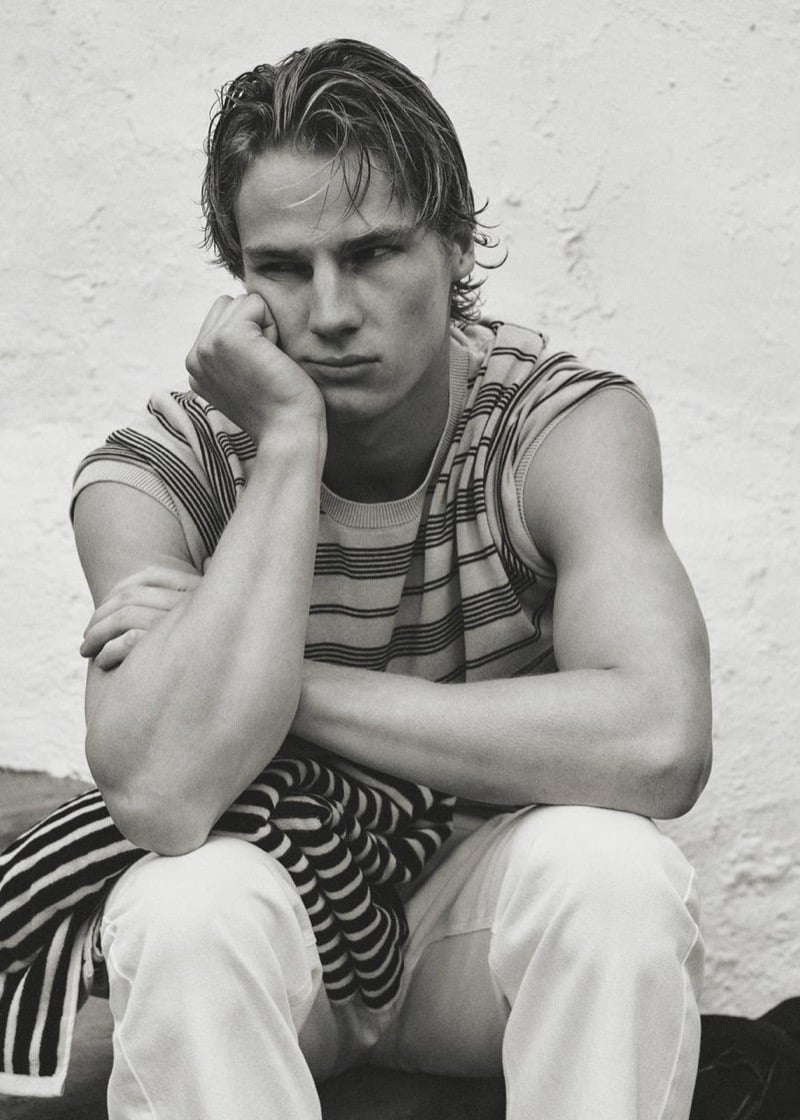 Henry Rank seated against a whitewashed wall in a horizontally striped t-shirt and cream trousers, a matching striped sweater knotted around the waist, wet swept-back hair, hand resting against his cheek