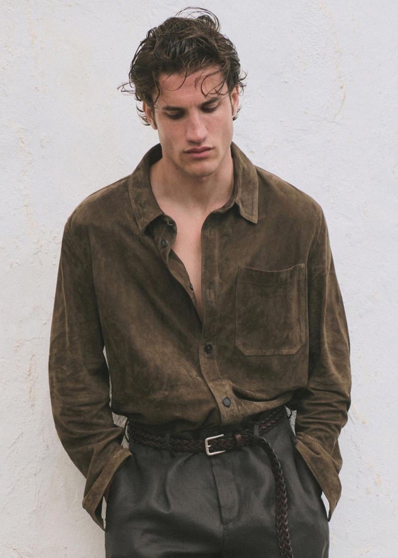 Max Knott in a chocolate brown suede shirt unbuttoned partway over olive linen pleated trousers with a braided leather belt, hands in pockets, wet curly hair, against a whitewashed wall