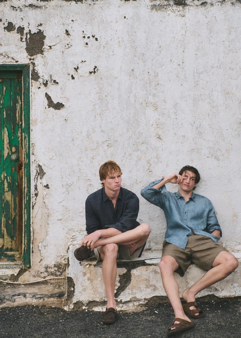 Henry Rank in a navy windowpane linen shirt with beige shorts and brown suede loafers and Max Knott in a faded blue linen shirt with tan linen shorts and brown suede slide sandals, both seated on a stone step in front of a weathered green wooden door
