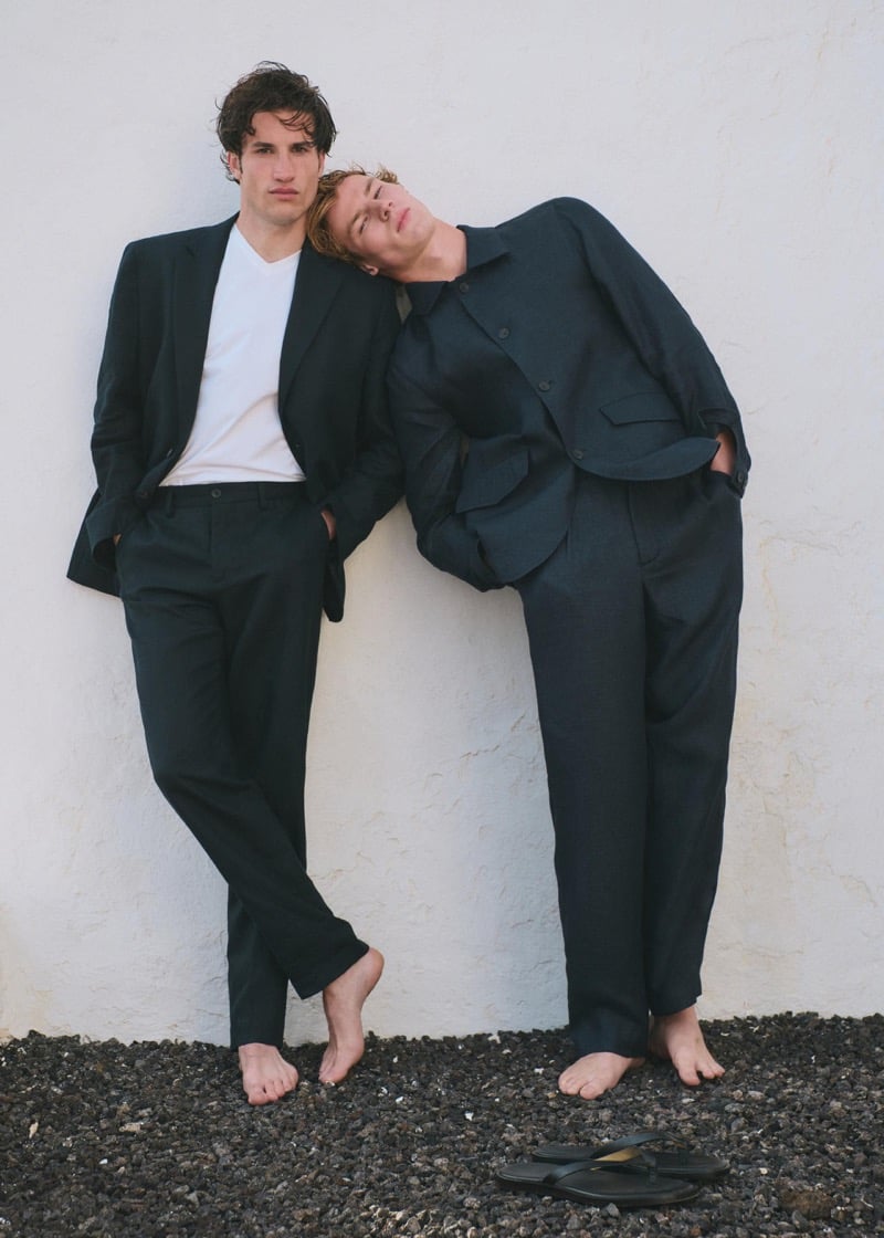 Max Knott in a dark navy linen suit with a white t-shirt and Henry Rank in an unbuttoned dark navy linen suit jacket and trousers, both leaning barefoot against a white plaster wall on black volcanic gravel, a pair of black thong sandals at their feet