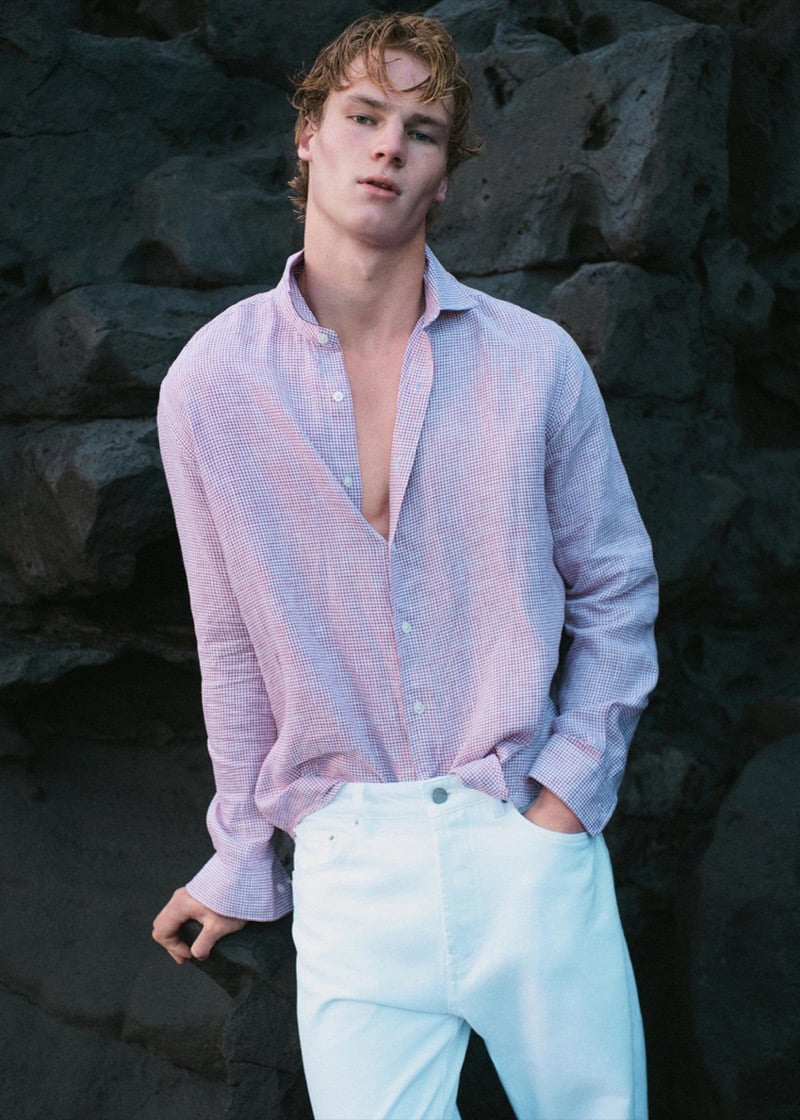 Henry Rank standing against a dark volcanic stone wall in a pink and pale blue micro-check linen shirt left open over the chest, paired with white denim jeans