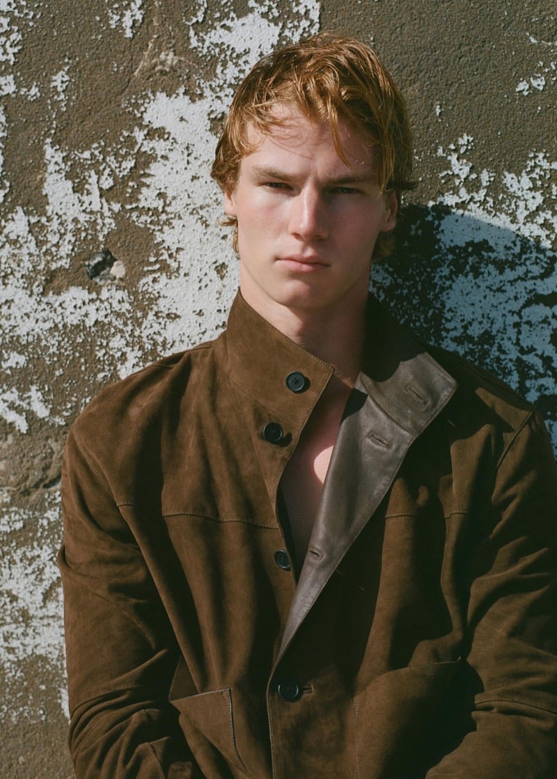 Henry Rank in close portrait wearing a brown suede mandarin-collar jacket with a leather panel collar partially buttoned, wet hair, against a sunlit peeling wall