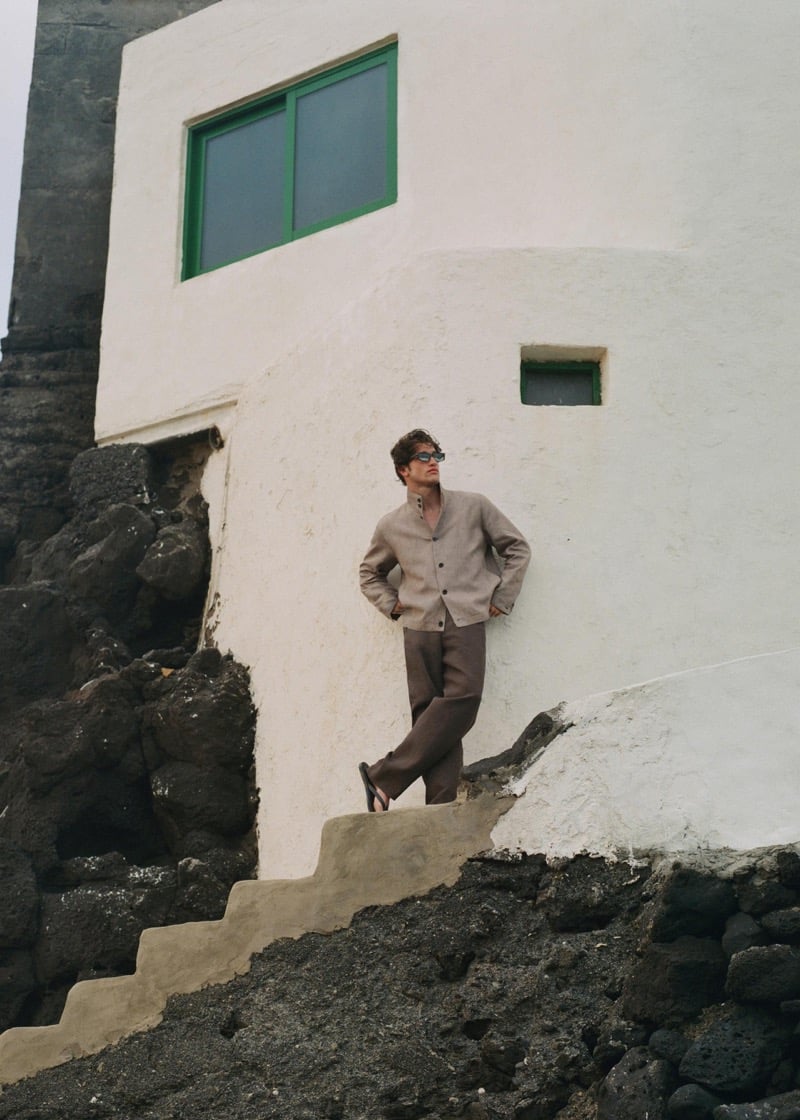 Max Knott standing on a cement stairway carved through black volcanic rock beside a whitewashed house with a green-trimmed window, wearing a flax linen cardigan, taupe trousers, black flip-flops, and dark sunglasses, hands behind his back