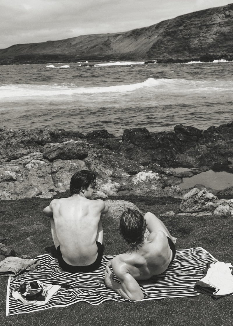 Max Knott and Henry Rank photographed from behind shirtless on a black and white striped beach towel on volcanic rocks at the shore, Max upright at left in dark swim shorts and Henry propped on his elbows at right, ocean and headlands beyond