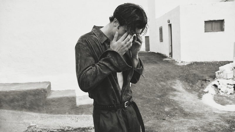 Max Knott photographed at half-length between two whitewashed buildings in a brown suede shirt unbuttoned to the chest over dark trousers cinched with a braided leather belt, hands pushing back wet hair