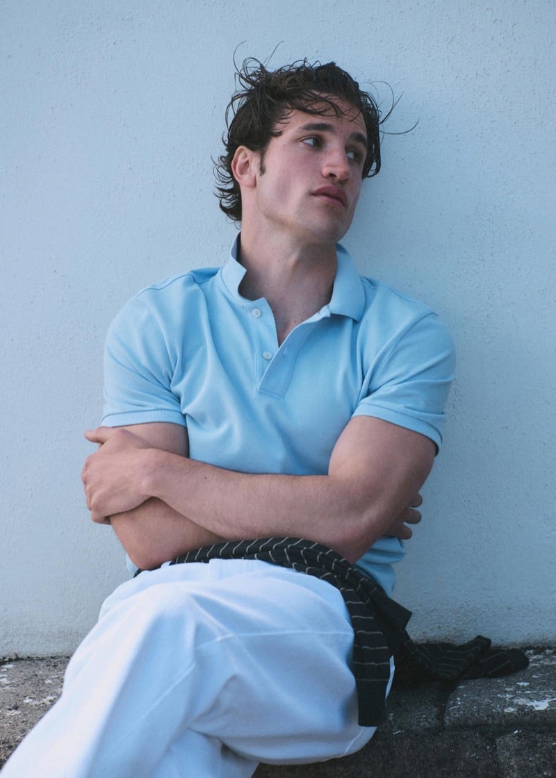 Max Knott seated against a pale blue plaster wall in a powder blue piqué polo and white trousers, a dark striped knit tied around the waist, arms folded, wet hair
