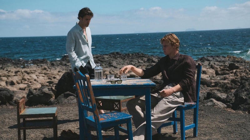 Max Knott standing in a white linen shirt and pale linen trousers and Henry Rank seated in a dark brown linen shirt and oatmeal linen trousers at a hand-painted blue wooden table set with a water carafe and glasses, volcanic coastline behind