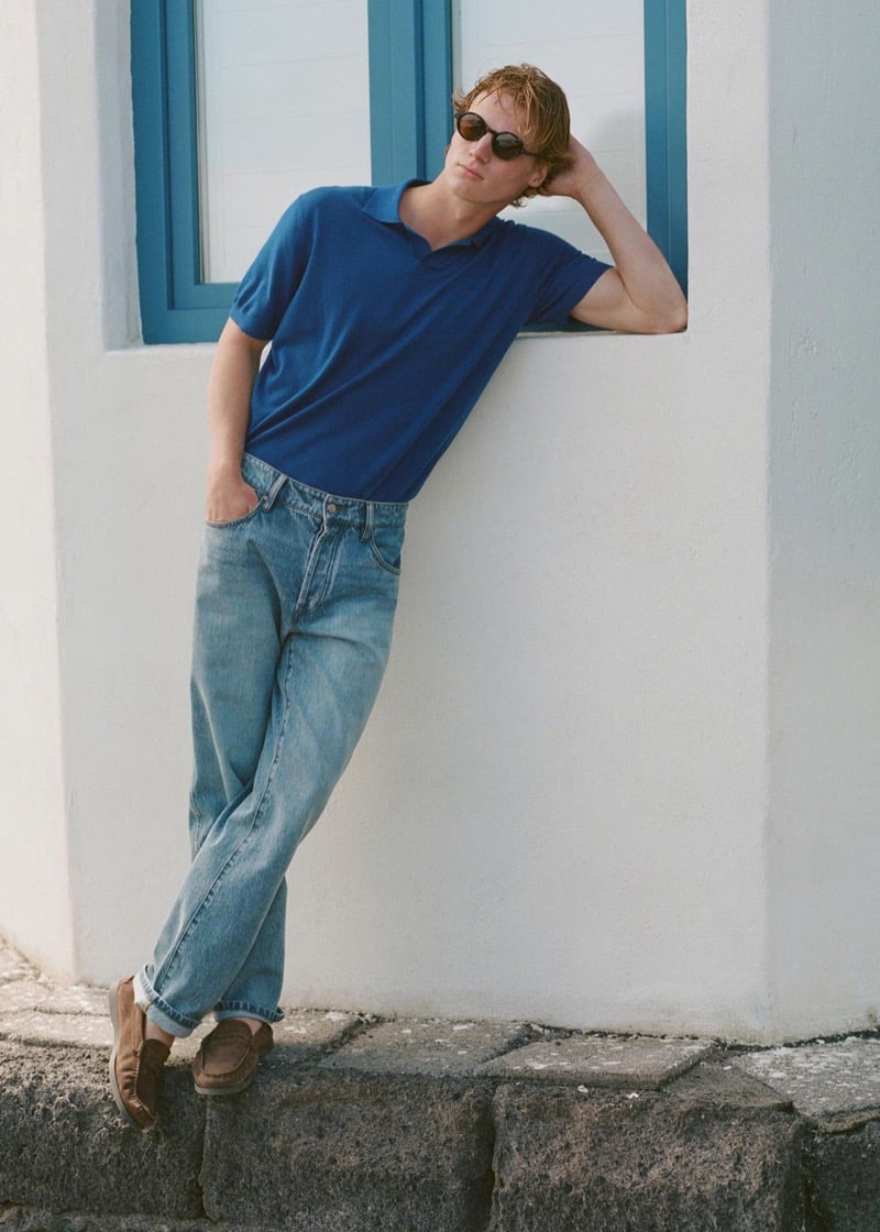 Henry Rank leaning against a white plaster wall with a blue-trimmed window, wearing a royal blue fine knit polo shirt, faded mid-blue straight-leg jeans, brown suede penny loafers, and dark round sunglasses