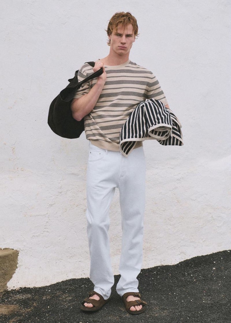 Henry Rank standing in front of a whitewashed wall in a cream and black horizontally striped t-shirt with white denim straight-leg jeans and brown suede slide sandals, a dark suede weekender bag over his shoulder and a navy and white striped towel folded over his arm