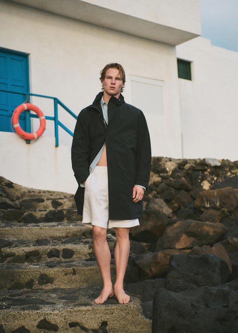 Henry Rank standing barefoot on volcanic stone steps beside a whitewashed fishing house with a blue handrail and orange life ring, wearing a long black parka over a pale sage shirt and white linen shorts