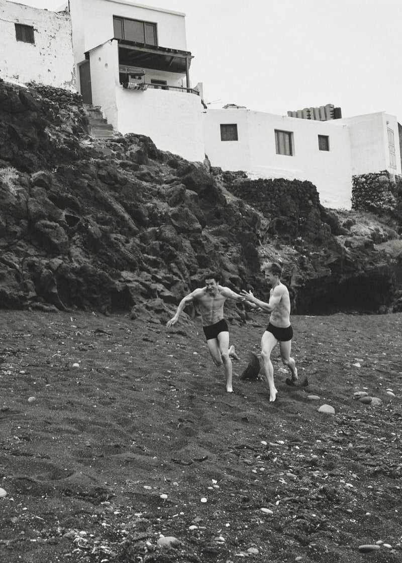 Max Knott and Henry Rank running shirtless across dark volcanic sand in matching black swim shorts, holding hands at full stride beneath a whitewashed cliffside village
