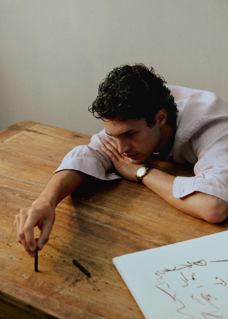 Francisco Henriques rests his head on a wooden table while sketching with a charcoal stick, wearing a linen shirt and a gold watch with a black leather strap