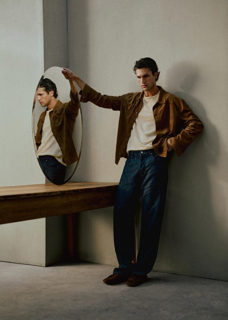 Francisco Henriques leans against a wall beside an oval floor mirror, wearing a tobacco brown suede shirt jacket over a cream knit tee with wide-leg dark indigo jeans and brown suede loafers