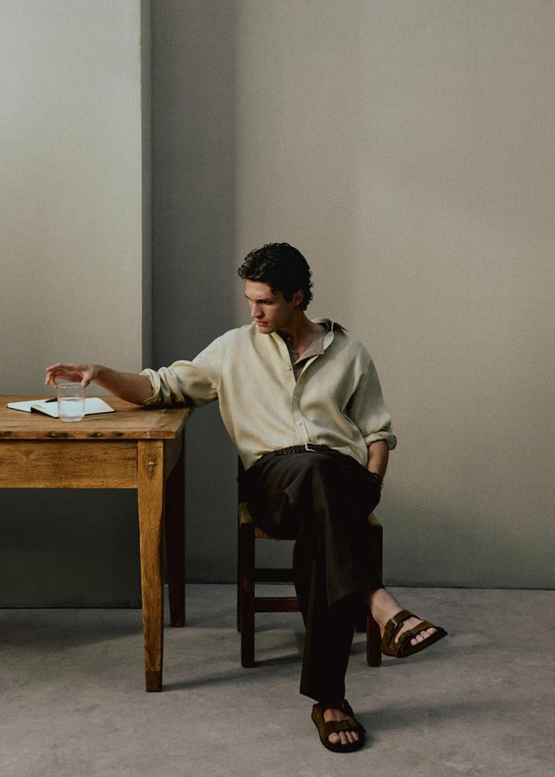 Francisco Henriques sits on a rush-seat wooden chair at a wooden table reaching for a glass of water, wearing an oversized cream linen shirt with dark brown trousers and brown suede slide sandals