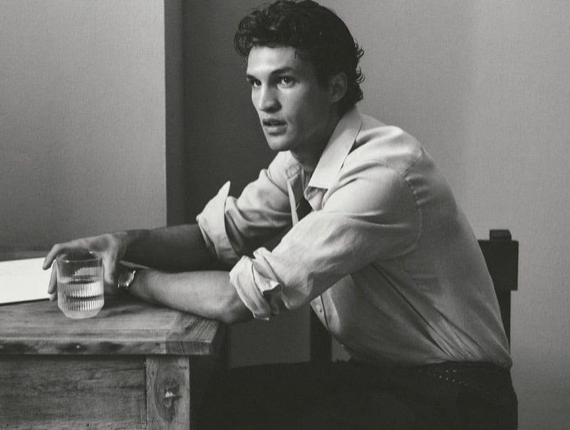 Black-and-white portrait of Francisco Henriques resting his forearms on a wooden table beside a glass of water, wearing a linen shirt with rolled sleeves