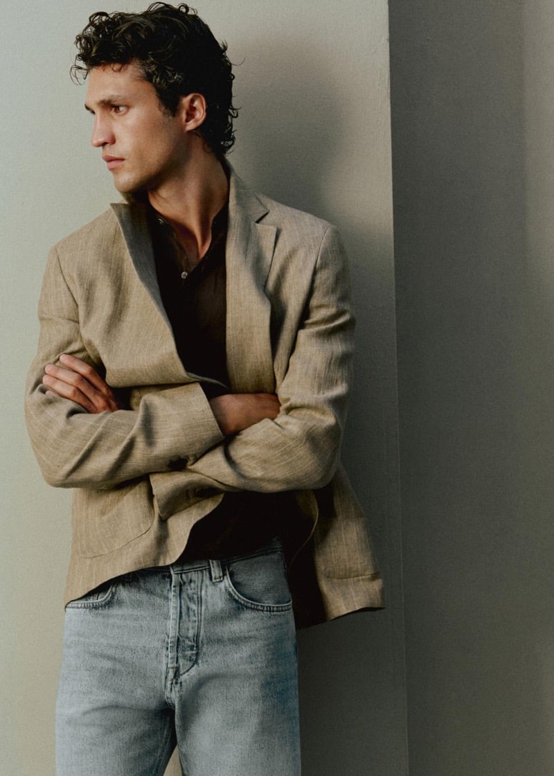Francisco Henriques leans against a wall with his arms crossed in an oatmeal linen blazer over a chocolate brown shirt with light wash straight-leg jeans