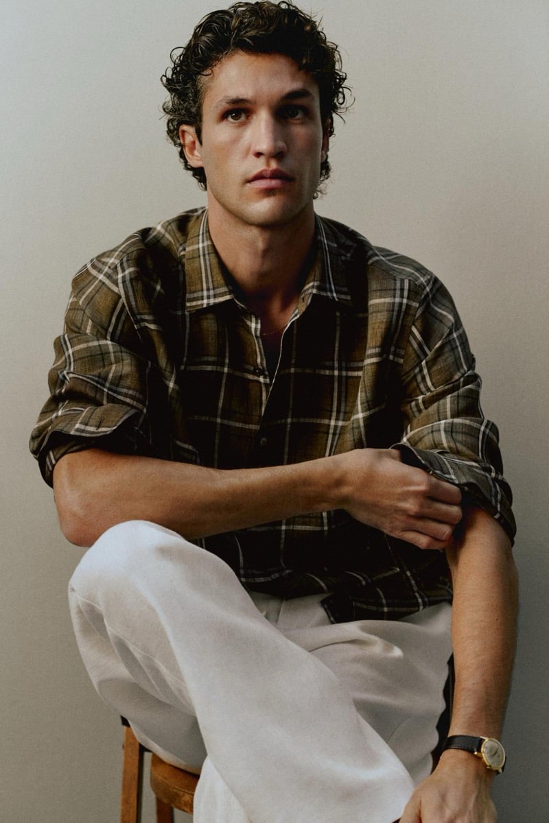 Francisco Henriques sits on a wooden stool in an olive plaid linen overshirt with rolled sleeves and white linen trousers, finished with a gold watch on a black leather strap