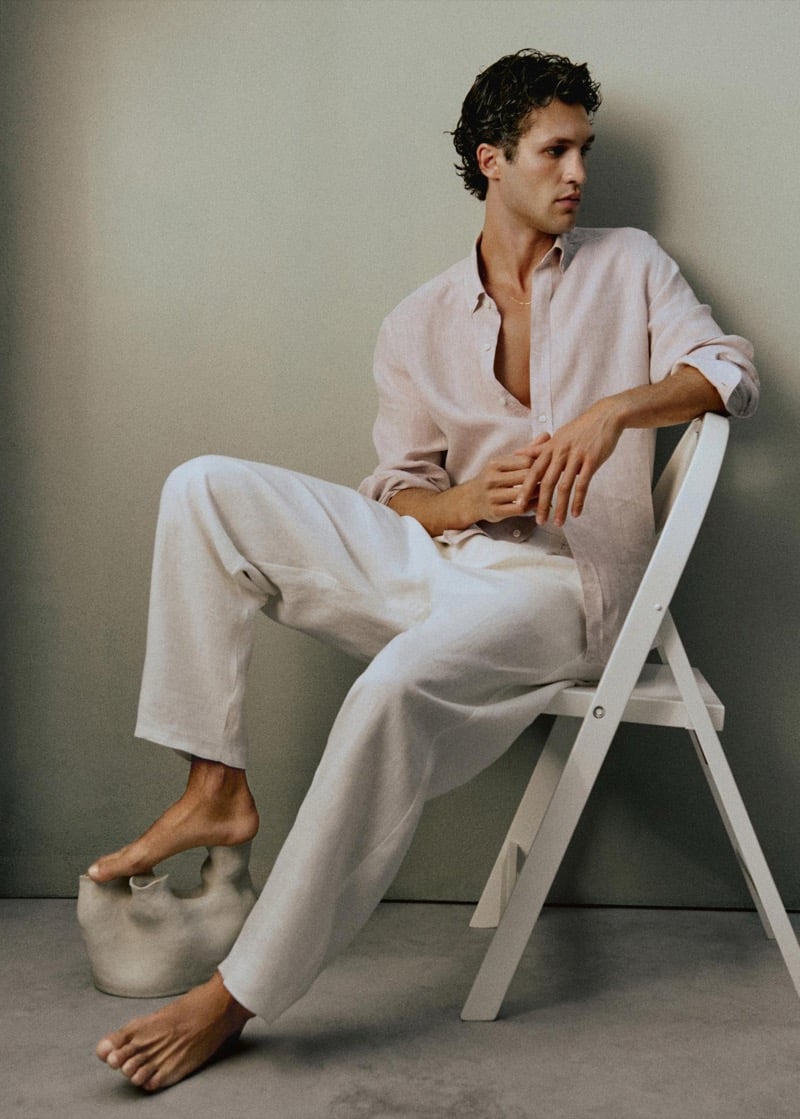 Francisco Henriques sits on a white folding chair with one foot resting on a ceramic vessel, wearing an unbuttoned dusty pink linen shirt with white linen trousers and a thin gold chain