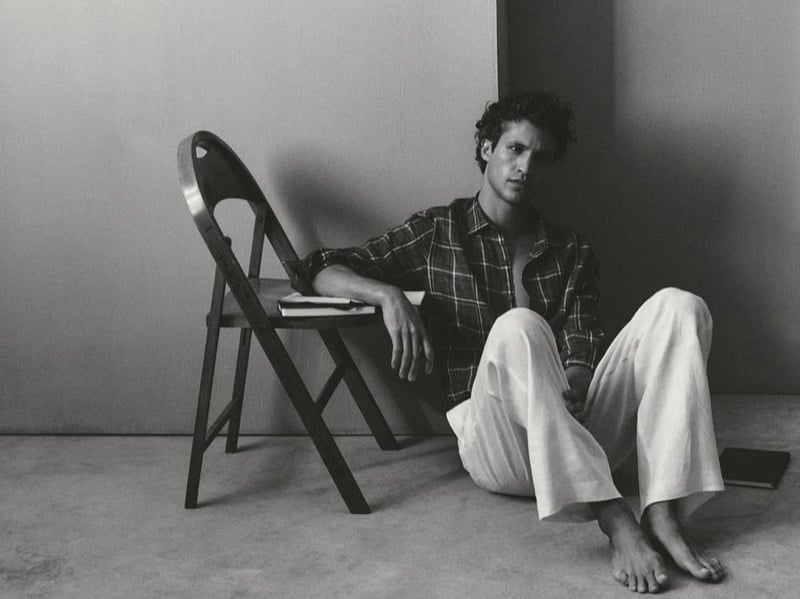Black-and-white image of Francisco Henriques sitting barefoot on the floor beside a wooden folding chair in an olive plaid shirt with cream linen wide-leg trousers