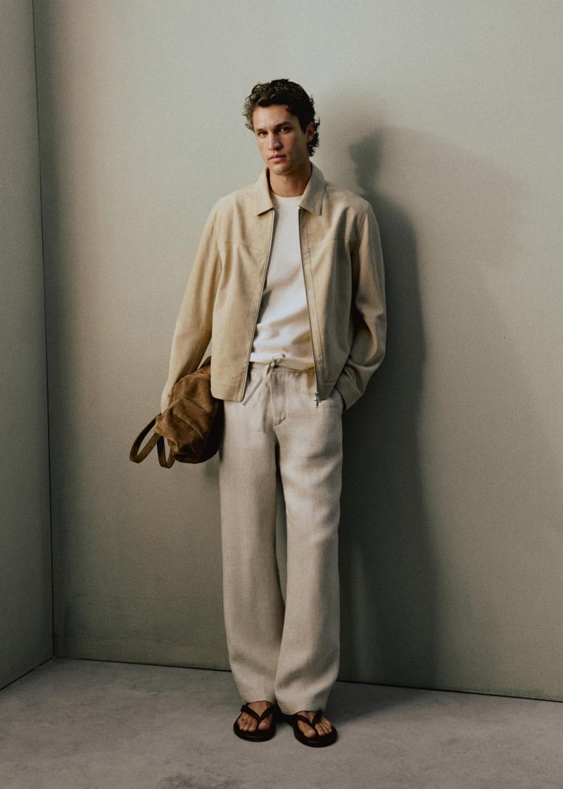 Francisco Henriques stands in a corner holding a tobacco suede tote bag, wearing a cream suede blouson jacket over a white tee with natural linen drawstring trousers and brown leather flip-flops